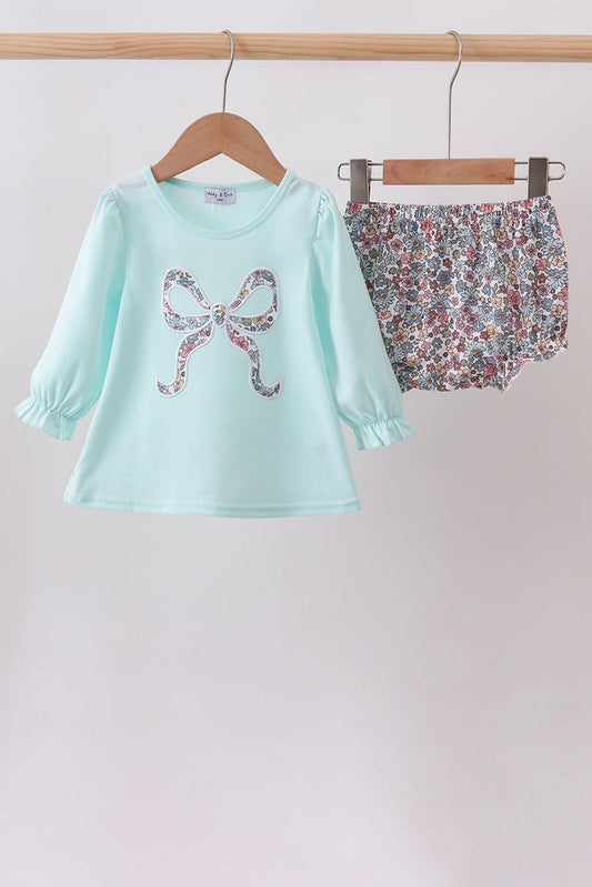 A two-piece girls' outfit hanging on wooden hangers from a wooden rod. The set includes a light mint green long-sleeve top with a bow-shaped floral print on the front, and matching floral print bloomers.